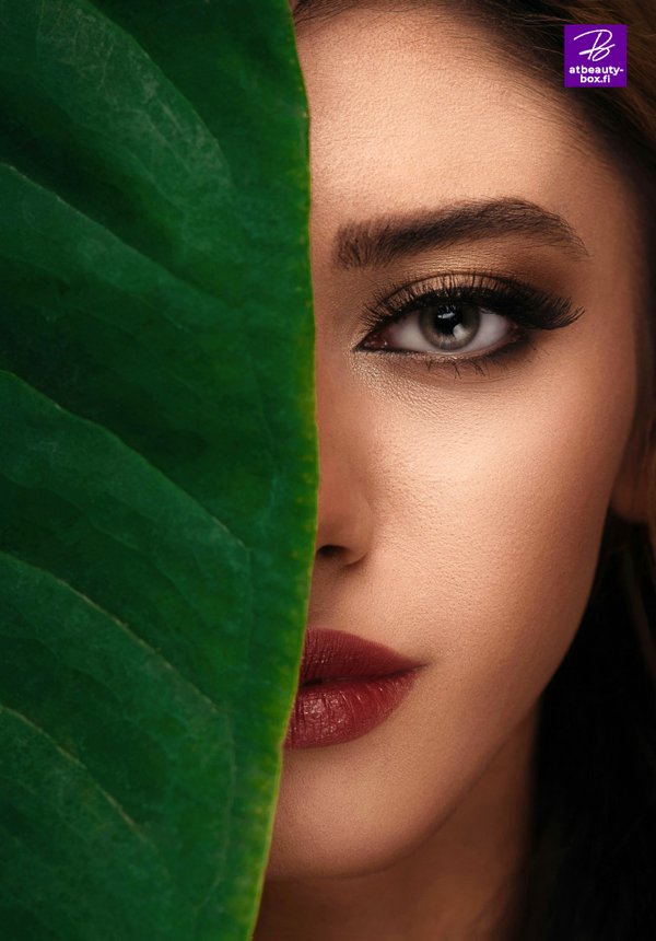A large dark green palm leaf covers half the face of a woman with make-up. The woman looks directly into the camera.